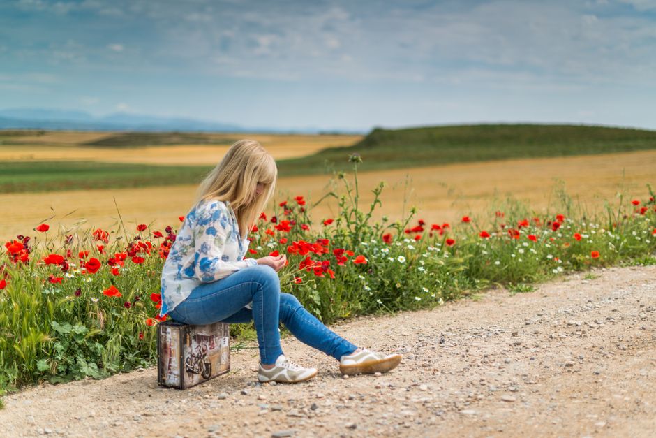 Mulher sentada em uma estrada rural, apreciando a paisagem de campo com flores silvestres e céu parcialmente nublado, transmitindo tranquilidade e contato com a natureza.
