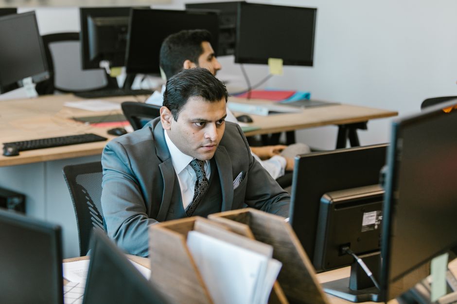 Homem de terno concentrado em frente ao computador em um escritório com múltimos monitores, sugerindo foco em análises ou trabalho intensivo.