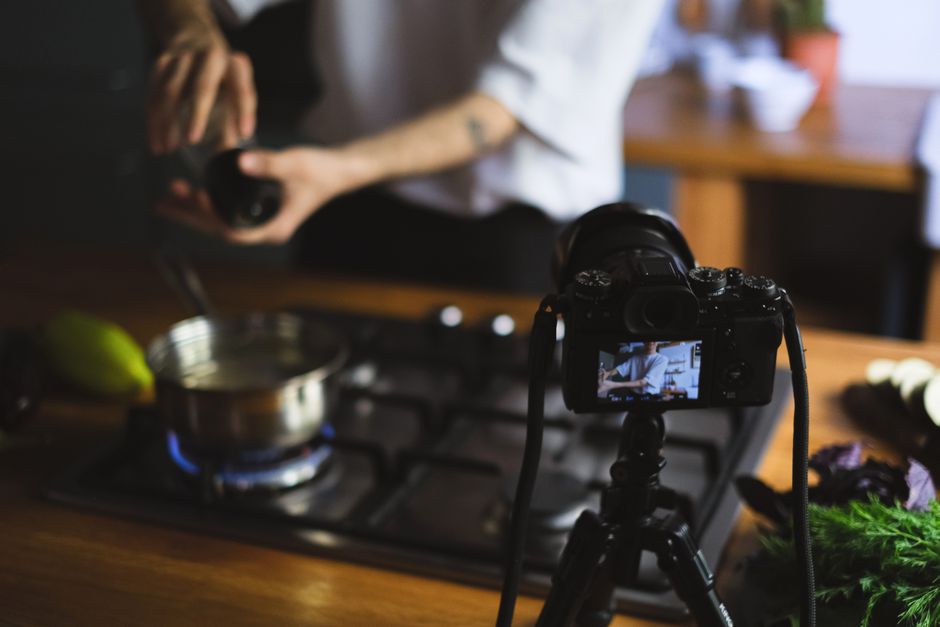 Imagem de uma pessoa gravando um vídeo na cozinha para um blog de culinária, com uma câmera posicionada em frente ao fogão aceso.