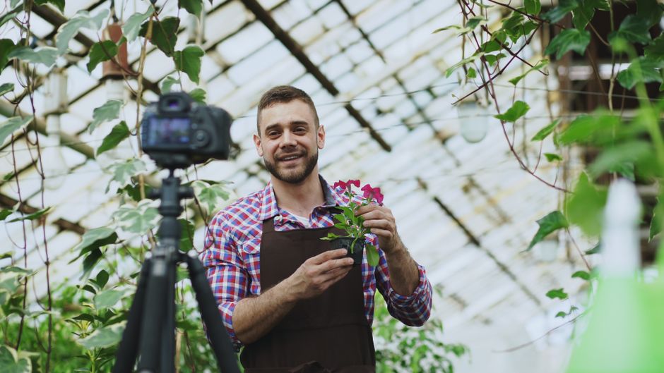 Homem realizando registro ou produção de conteúdo em ambiente de estufa ou viveiro de plantas, com foco em horticultura ou agricultura sustentável.