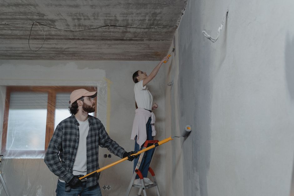 Pessoa pintando uma parede de uma casa em reforma, enquanto outro acompanha com uma ferramenta de pintura.