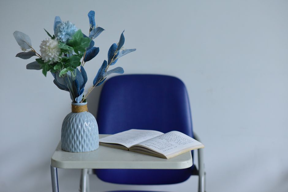 Imagem de uma cadeira azul com uma mesa ao lado, contendo um livro aberto e um vaso com flores artificiais, ideal para ilustrar temas de leitura, organização ou decoração de ambientes.