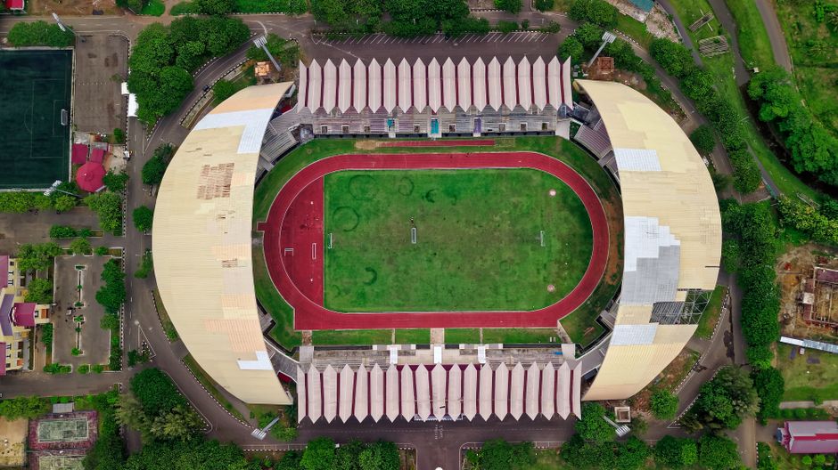 Vista aérea de um estádio esportivo com campo de futebol e pista de atletismo, rodeado por árvores e áreas urbanas.