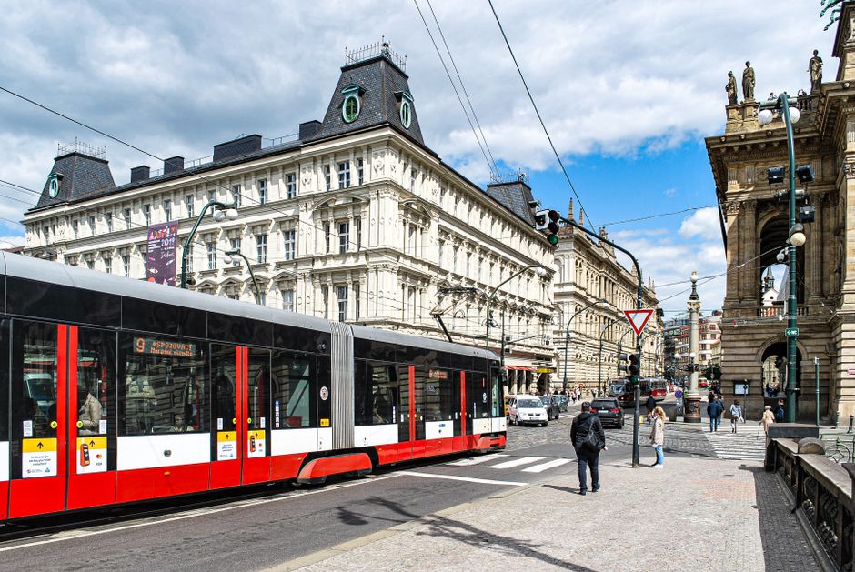 Imagem de uma rua urbana com um bonde moderno passando ao lado de edifícios históricos, em uma cidade movimentada, sob céu parcialmente nublado.