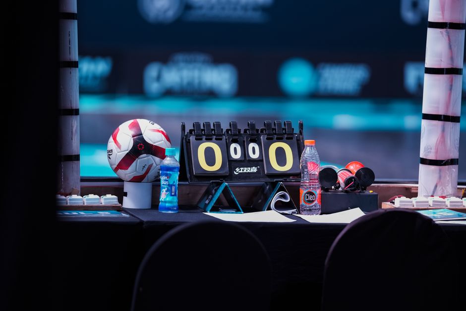 Mesa de controle com equipamentos utilizados em eventos esportivos, destacando cronômetro, bola de futebol, garrafas de água e fones de ouvido, em ambiente de competição.