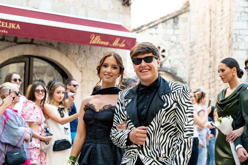 Casal elegante sorrindo em um evento ao ar livre, cercado por pessoas que parecem participar de uma celebração ou festa social.