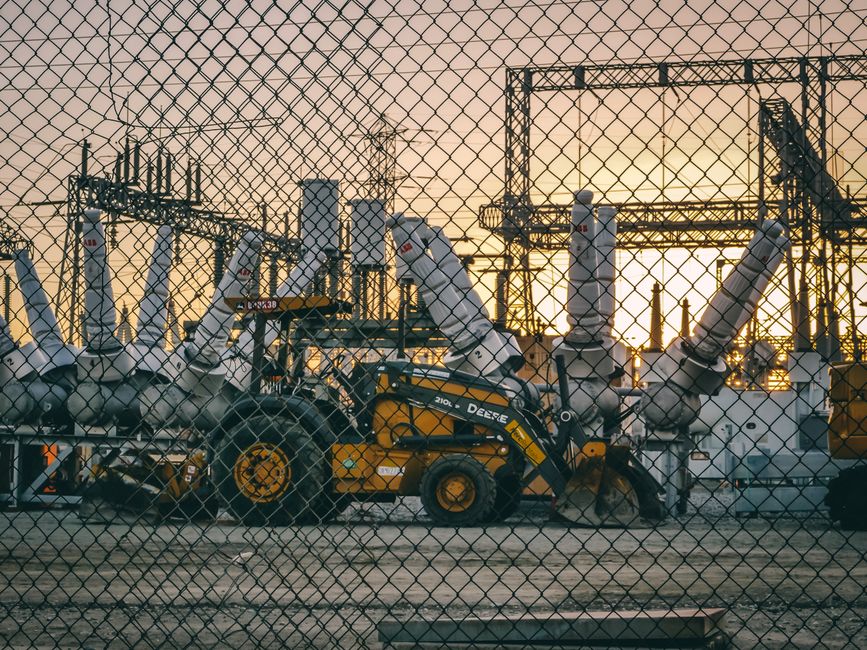 Imagem de uma usina de energia elétrica industrial mostrada através de uma cerca, com foco na infraestrutura de geração de energia ao entardecer.