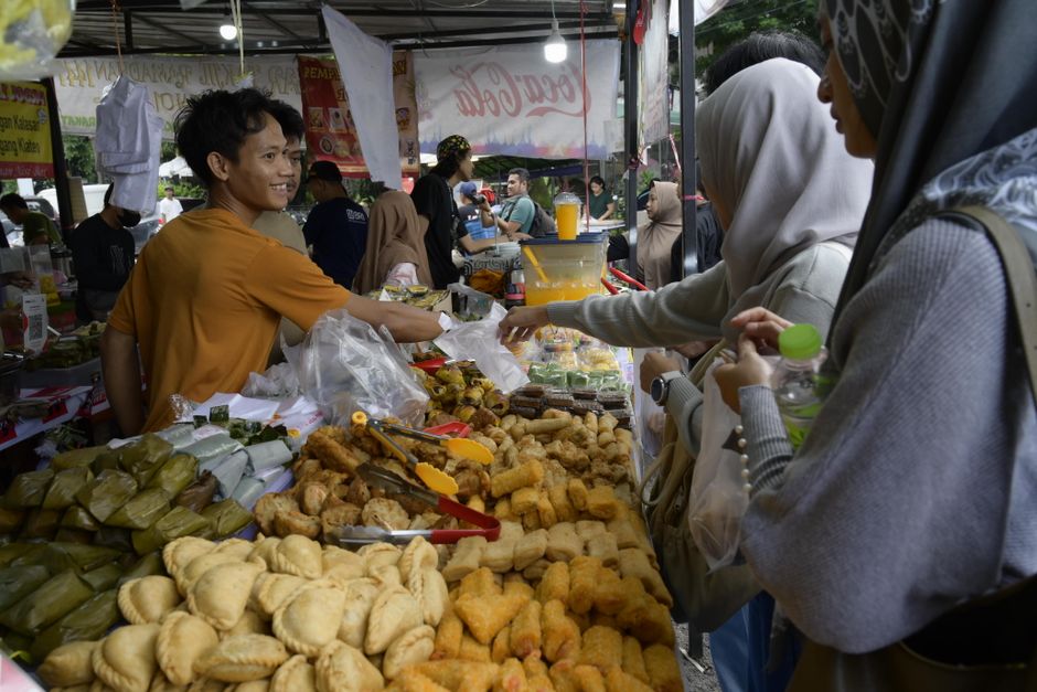 Vendedora atendendo clientes em feira de alimentos ao ar livre, com variedade de comidas tradicionais expostas na bancada.