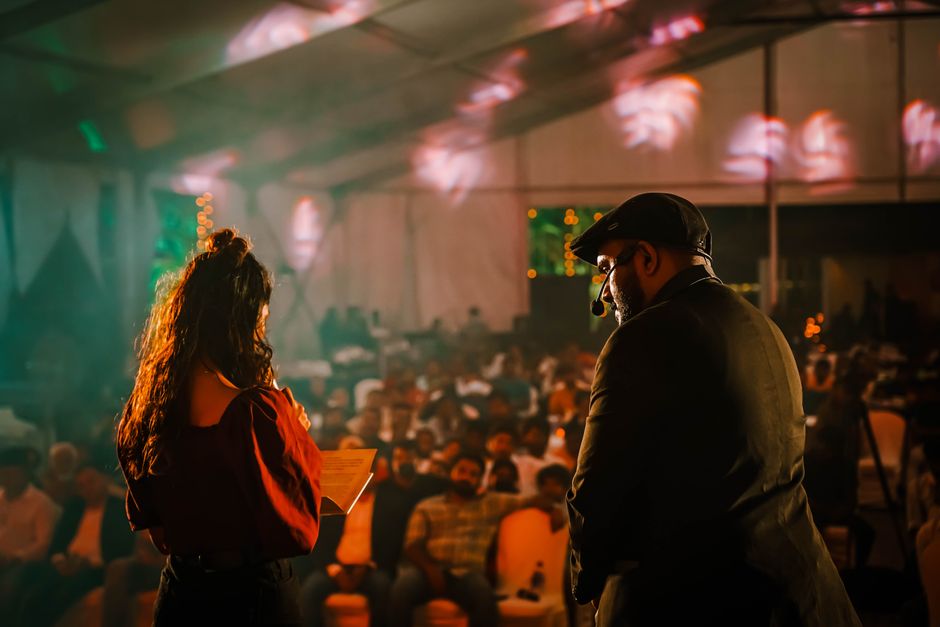 Imagem de um apresentador e uma apresentadora em um palco durante um evento ou palestra com público ao fundo.
