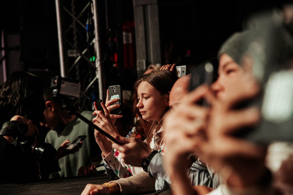 Grupo de pessoas em uma conferência ou evento, focando na captura de momento com seus celulares, representando interesse e envolvimento em uma atividade ao vivo.