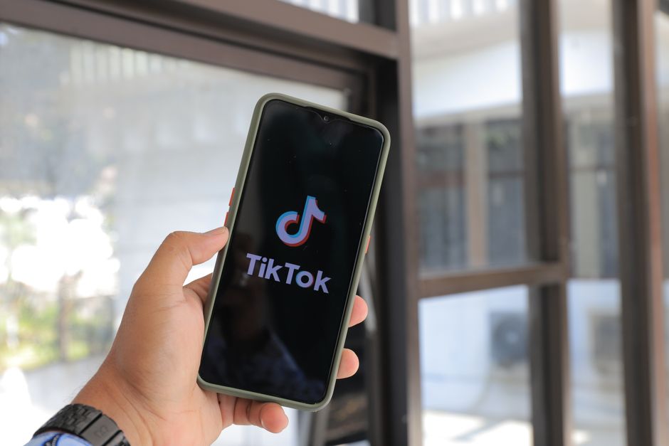 Pessoa segurando um smartphone exibindo o logo do TikTok, com ambiente de escritório ao fundo, evidenciando o uso de redes sociais no trabalho.