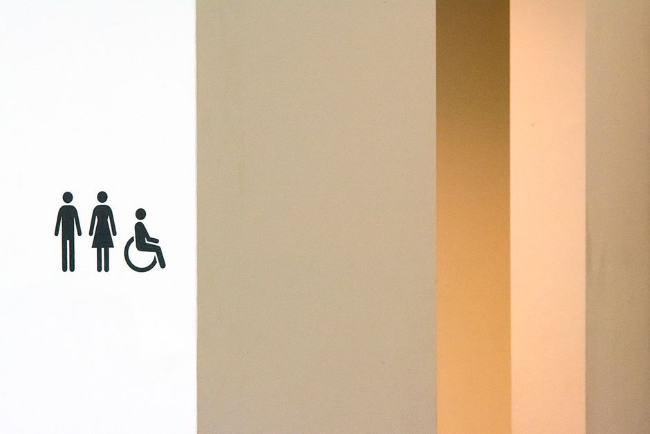 Sinalização de acessibilidade indicando o percurso para o banheiro com simbolismos de gênero e cadeira de rodas.