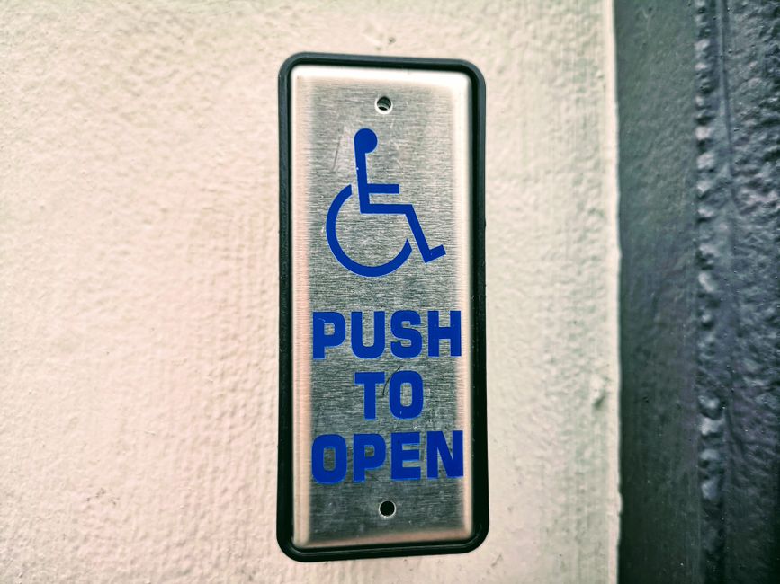 Placa de acessibilidade indicando necessidade de pressionar para abrir uma porta, destacando a inclusão e acessibilidade para pessoas com deficiência.